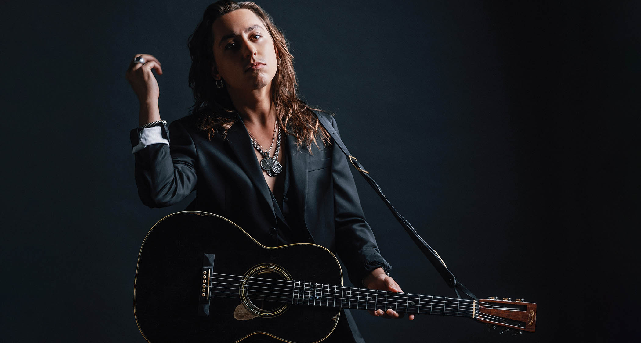 Jake Kiszka wears a dark suit, no shirt, and is photographed with his custom made Martin acoustic.